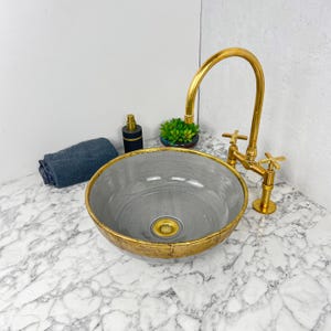 Puede incluir: Un lavabo de baño de cerámica gris con un borde dorado y un grifo dorado. El lavabo está sobre una encimera de mármol blanco.