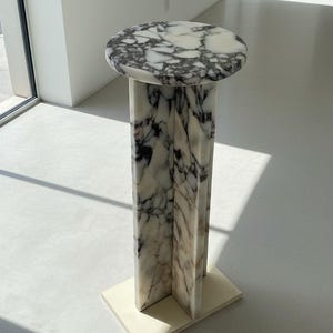 May include: A tall pedestal stand crafted from white marble with prominent black veining. The design features a circular top surface supported by a central column composed of three intersecting vertical panels, resting on a flat rectangular base.