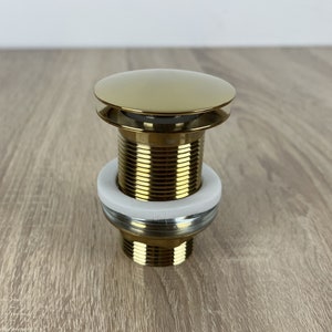 Puede incluir: Un tapón de desagüe emergente dorado para un lavabo de baño. El tapón tiene una parte superior redonda y una base roscada. Incluye una junta de goma blanca.