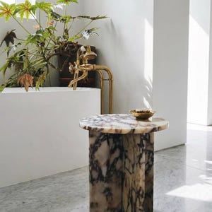 May include: A cylindrical side table crafted from white and dark purple veined marble with a smooth circular top. A small gold-toned bowl sits on the surface. The table is positioned in a bright room near a white bathtub with a brass faucet fixture.