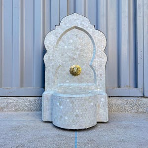 Może przedstawiać: Biała fontanna ścienna wykonana z mozaikowych płytek z mosiężnym wylotem w kształcie głowy lwa. Fontanna ma tradycyjny marokański design.