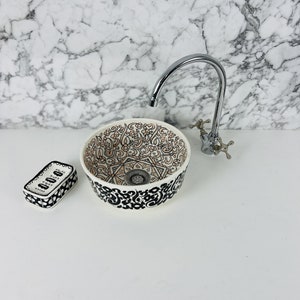 May include: A white ceramic sink with a black and peach floral design. The sink is round and has a drain in the centre. A chrome faucet is mounted to the wall above the sink.
