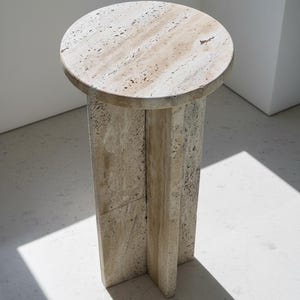 Op de afbeelding: Een cilindrische bijzettafel gemaakt van beige travertijnsteen, met een rond tafelblad en een onderstel bestaande uit drie kruisende verticale panelen die een kruisvormige steunstructuur vormen met een poreuze, natuurlijk getextureerde afwerking.