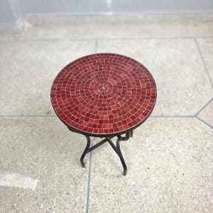 Könnte beinhalten: Ein kleiner, runder Tisch mit einer Tischplatte aus roten Mosaikfliesen und einem schwarzen Metallgestell. Der Tisch hat ein schlichtes, elegantes Design.