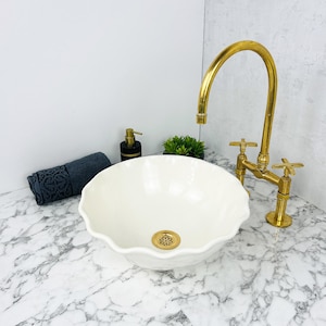 May include: White ceramic bathroom sink with a wavy edge, a gold drain, and a gold faucet with two handles. The sink is on a white marble countertop.