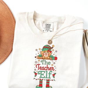 May include: Cream-colored t-shirt featuring a cartoon elf holding a pencil, with the text "The Teacher Elf" in red and green. The elf wears a green hat and red and green striped legs. A gold necklace is draped over the shirt.