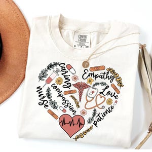 May include: Cream-colored t-shirt with a heart-shaped design featuring medical symbols, flowers, and words like "nurse," "caring," and "empathy." A gold necklace and a brown hat are visible. The shirt is from Comfort Colors.