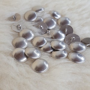 Könnte beinhalten: Ein Haufen silberner Metallknöpfe auf einer weißen, flauschigen Oberfläche.