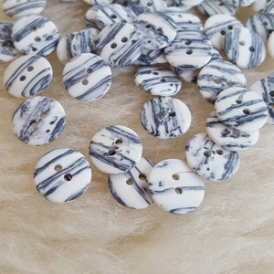 Könnte beinhalten: Eine Sammlung von weißen und schwarzen gestreiften Knöpfen mit jeweils zwei Löchern. Die Knöpfe sind auf einer weißen, flauschigen Oberfläche verstreut.