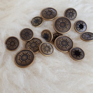 Könnte beinhalten: Eine Sammlung antiker, bronzefarbener Knöpfe mit kreisförmigem Design. Die Knöpfe sind auf einer weißen, flauschigen Oberfläche verstreut. Die Knöpfe haben ein detailliertes Muster an den Rändern und eine dunkle Mitte.