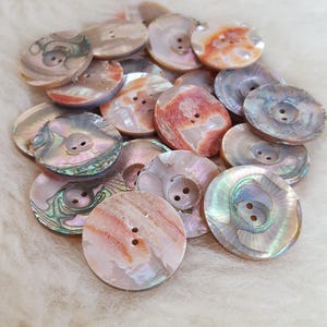 Könnte beinhalten: Eine Sammlung von irisierenden Perlmuttknöpfen in verschiedenen Schattierungen von Rosa, Weiß und Braun. Die Knöpfe haben eine glatte, abgerundete Oberfläche und ein Zwei-Loch-Design.
