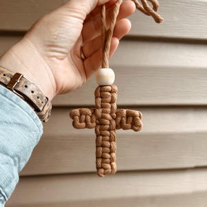 May include: A macrame cross ornament crafted from brown cord, featuring a single light-colored wooden bead at the top. The cross is suspended by a matching brown cord loop, showcasing intricate knotting patterns throughout the entire structure.