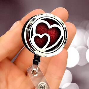 Puede incluir: Un portatarjetas redondo plateado con un diseño de corazón rojo. El portatarjetas tiene un borde negro y una correa de plástico transparente con cierre a presión. El diseño presenta dos corazones, uno dentro del otro, con un fondo rojo texturizado.