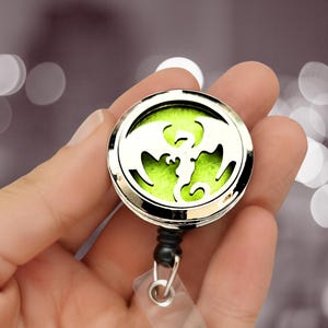 Puede incluir: Un carrete de insignia redondo plateado con un diseño de dragón. El dragón está recortado, revelando un fondo verde brillante. El carrete tiene un cordón negro y una correa de plástico transparente.