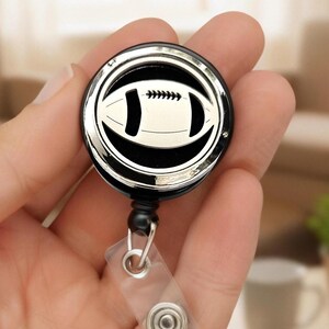 Puede incluir: Un carrete retráctil para insignia negro y plateado con un diseño de balón de fútbol. El carrete tiene forma circular con un balón de fútbol plateado brillante en el centro, rodeado por un fondo negro y un borde plateado. Se adjunta una correa de plástico transparente y un clip de metal.