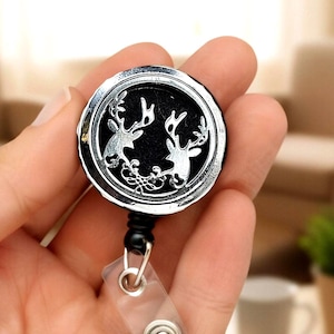 Puede incluir: Un carrete retráctil de insignia plateado y negro con dos siluetas de ciervos enfrentadas. El carrete de insignia tiene una correa de plástico transparente y un cordón negro. El diseño es circular y tiene un borde decorativo.