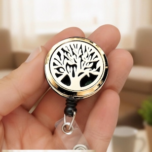 May include: A circular gold-toned badge reel featuring a cut-out tree of life design on the front face. The metallic housing has a polished finish, and a clear plastic strap with a metal snap button extends from the bottom of the black plastic connector.