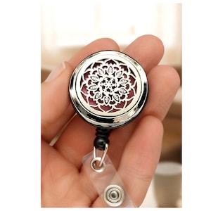 Puede incluir: Un carrete retráctil para insignias plateado y negro con un diseño floral decorativo. El centro presenta un fondo burdeos. El carrete tiene una correa de plástico transparente y un clip metálico.