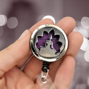 Puede incluir: Un portatarjetas plateado con un diseño redondo. Presenta un elemento morado con forma de flor y una figura sentada. El portatarjetas tiene un cordón retráctil y un soporte transparente para tarjetas.