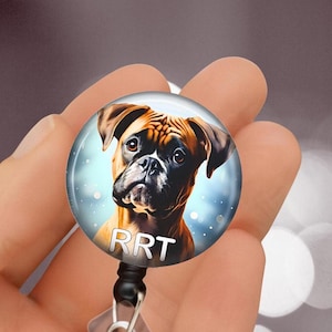 May include: A round retractable badge reel with a Boxer dog portrait. The dog has brown and white fur with dark eyes and ears. The letters "RRT" are printed in white. The reel has a black cord and a metal clip.