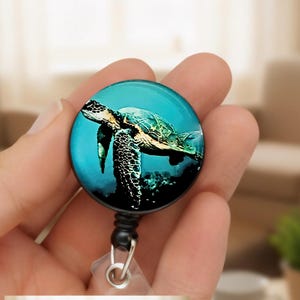May include: A circular badge reel featuring a high-resolution image of a sea turtle swimming in deep blue water. The turtle has a patterned shell and flippers, set against a dark, aquatic background. The reel has a black frame and a metal clip.