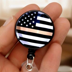May include: A round badge reel featuring a design of the American flag with a thin gold line. The badge reel has a black border and a clear plastic strap for attaching to clothing. The design includes white stars on a blue background.