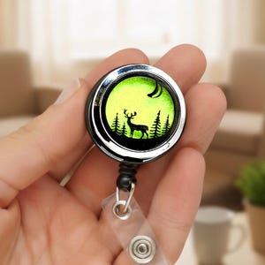 Puede incluir: Un carrete retráctil para insignia negro y plateado con una escena de la naturaleza. El diseño presenta una silueta de un ciervo, árboles y una luna creciente sobre un fondo verde y amarillo. El carrete tiene una correa de plástico transparente.