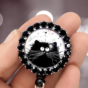 May include: A circular badge reel featuring a graphic illustration of a fluffy black cat with wide white eyes and long whiskers against a white background. The badge is framed by a border of small, faceted black stones set in silver metal.