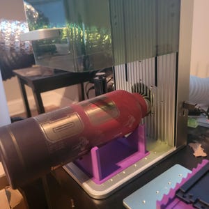 以下が含まれることがあります： 赤と金のウォーターボトルがレーザーマシンで彫刻されています。ボトルは紫色のプラスチックスタンドで固定されています。マシンには透明なプラスチックカバーと銀色の金属フレームが付いています。