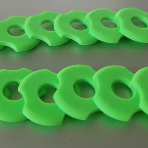 Puede incluir: Doce objetos verdes brillantes en forma de anillo con un diseño de borde festoneado único. Dispuestos en dos filas paralelas de seis, estos objetos sugieren un conjunto o colección. Cada objeto tiene un agujero central.