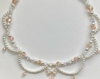 May include: A delicate white and pink pearl necklace with a silver bow and flower charm. The necklace has a layered design with multiple strands of pearls.