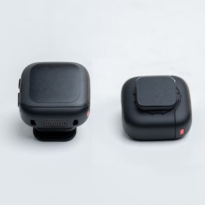 May include: Two black, square-shaped devices are displayed against a white background. One device has a rounded top and a small red dot on the side. The other device has a flat top with a smaller square on top and a red dot.