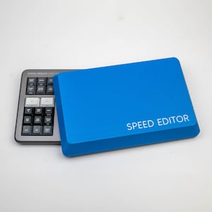 May include: A blue external hard drive with the text "SPEED EDITOR" printed on the front. The hard drive is next to a grey keyboard with white keys labelled with functions such as "CUT", "SLIP", "TRANS", and "MARK".