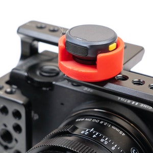 Può includere: Una fotocamera nera con un accessorio rosso e giallo attaccato alla parte superiore. L'obiettivo della fotocamera ha un anello nero con segni bianchi e il testo "45mm 1:2.8 DG DN".