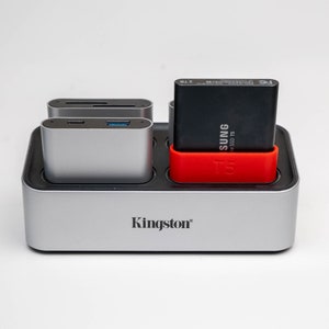 May include: A silver Kingston hard drive docking station with a black Samsung T5 SSD drive inserted. The docking station has a red rubber sleeve around the SSD drive.