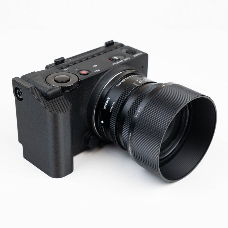 May include: A black camera with a large lens attached. The camera has a black hand grip and a red button on the top. The lens is a Sigma 45mm f/2.8 DG DN Contemporary lens.