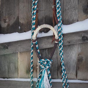 May include: A horse halter and lead rope set featuring a braided pattern in teal, white, and black. The halter includes a white rawhide noseband, dark brown leather accents, and a decorative tassel at the base of the chin strap.