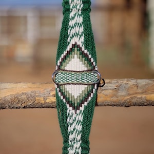 May include: A green and white woven horse cinch with a diamond pattern. The cinch features a metal buckle and rings. The design includes shades of green, white, and brown. The cinch is displayed against a blurred outdoor background.
