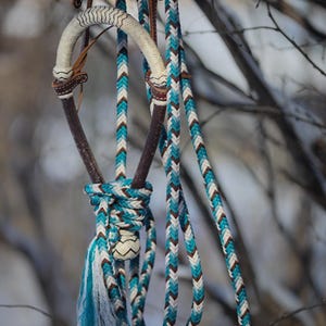 May include: A horse bridle featuring a braided rope design in turquoise, white, and dark brown. The headstall includes a curved, cream-colored braided noseband and dark brown leather accents, with long braided reins hanging down in a knot.