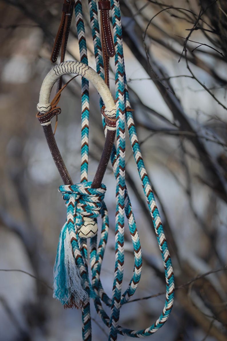 May include: A horse bridle featuring a braided rope design in turquoise, white, and dark brown. The headstall includes a curved, cream-colored braided noseband and dark brown leather accents, with long braided reins hanging down in a knot.