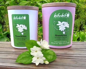 May include: Two soy wax candles in glass jars with wooden lids. The candles are labeled "flowerlicious sampaguita (jasmine) scented candle". The candle on the left is white and the candle on the right is pink. Both candles have a green label with a white flower illustration.