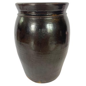 May include: A dark brown, cylindrical stoneware crock with a glossy, reflective glaze. The vessel features a wide, slightly flared rim and a thick, rounded collar at the top. The surface has a smooth, mottled texture with subtle variations in color.