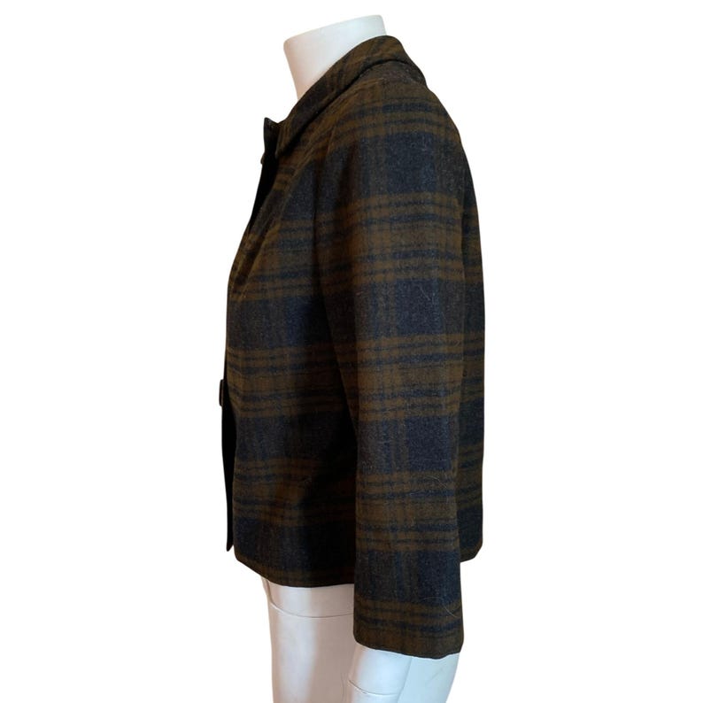 May include: A long-sleeved blazer featuring a dark charcoal gray and olive green plaid pattern. The garment has a structured collar, a single-breasted front closure with visible buttons, and a straight hemline, displayed on a white mannequin.