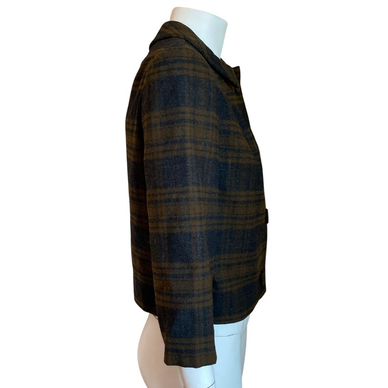 May include: A cropped jacket featuring a dark brown and charcoal gray plaid pattern. The garment has a collared neckline, long sleeves, and a button-front closure. The fabric appears to have a textured, wool-like finish, and the jacket is displayed on a mannequin to show its structure and fit.