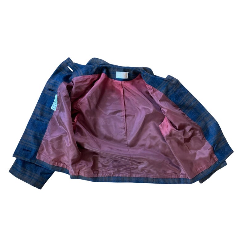 May include: A blue and brown plaid jacket featuring a solid mauve-colored interior lining. The garment includes a white brand label at the inner neckline and a small white rectangular tag attached to the inner side seam of the left front panel.
