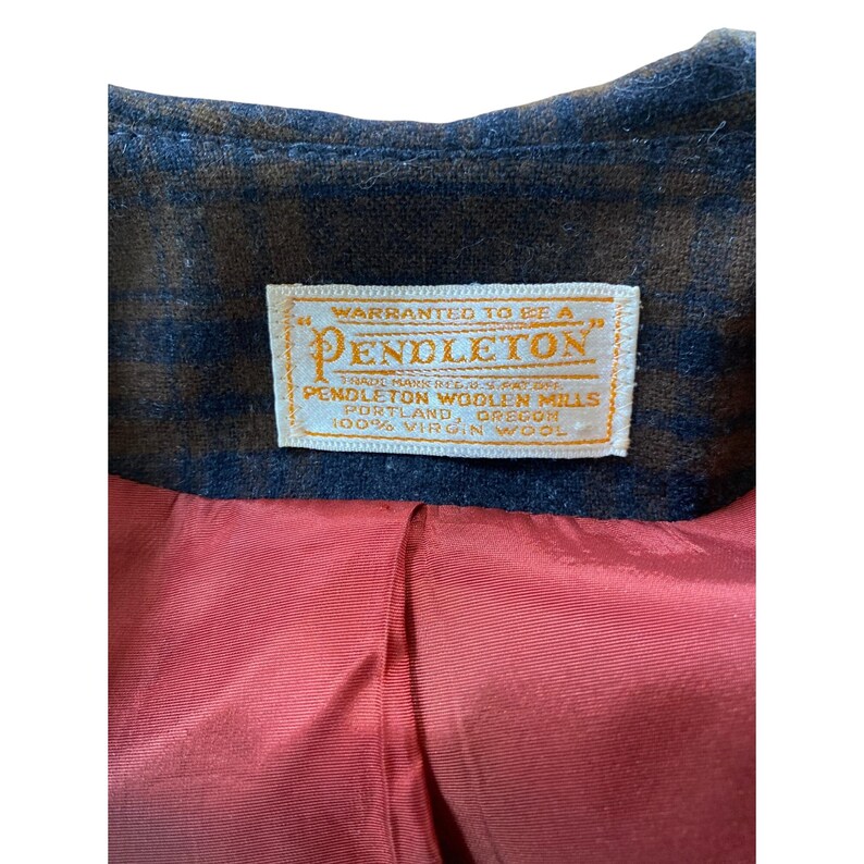 May include: A brown and navy blue plaid wool jacket with a red lining. A white rectangular label is sewn into the collar area, featuring the text: Warranted to be a Pendleton, Pendleton Woolen Mills, Portland, Oregon, 100% Virgin Wool.