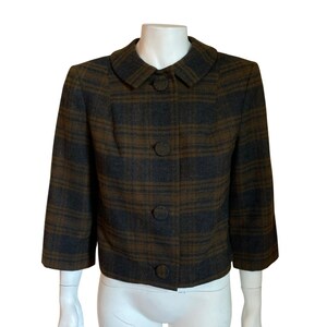 May include: A cropped jacket featuring a dark brown and charcoal gray plaid pattern. The garment has a rounded collar, three-quarter length sleeves, and a front button closure with four large, fabric-covered buttons in a matching plaid design.