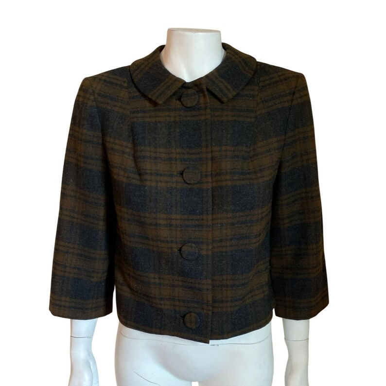 May include: A cropped jacket featuring a dark brown and charcoal gray plaid pattern. The garment has a rounded collar, three-quarter length sleeves, and a front button closure with four large, fabric-covered buttons in a matching plaid design.