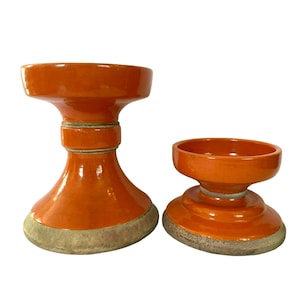 May include: Two orange ceramic candle holders with a wide base and a narrow middle section. The larger holder is taller than the smaller holder.