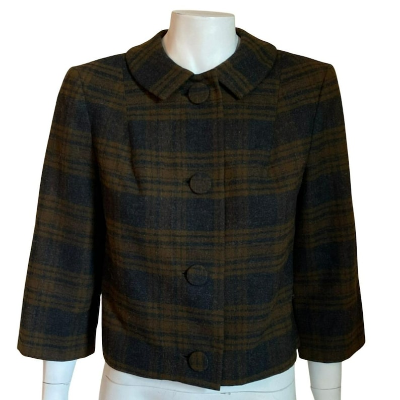 May include: A cropped jacket featuring a dark brown and charcoal gray plaid pattern. The garment has a rounded collar, three-quarter length sleeves, and a front button closure with four large, matching fabric-covered circular buttons.